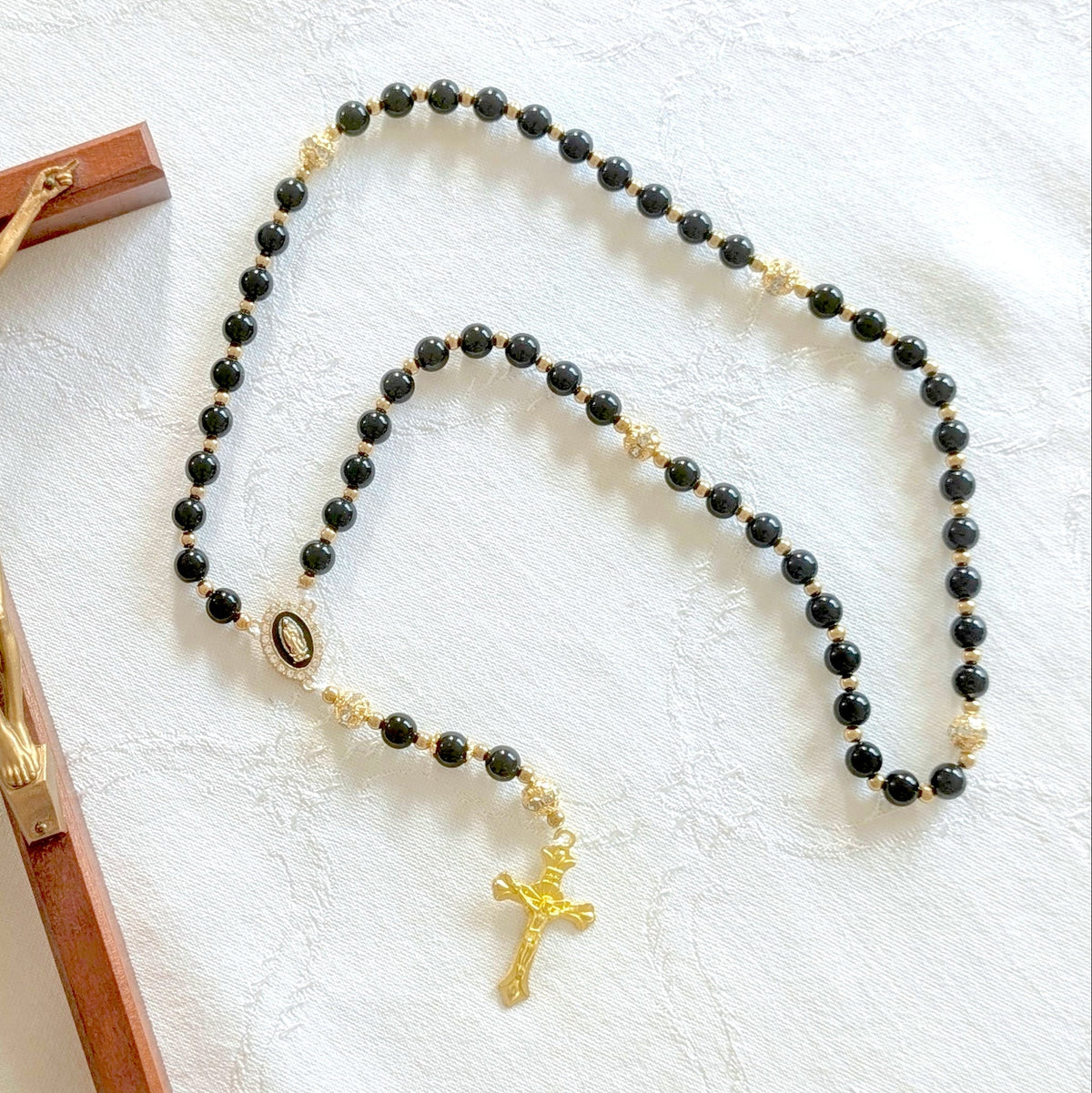 A Catholic rosary with black beads and gold accents, displayed next to a wooden crucifix with a gold metal corpus on a textured white fabric background.